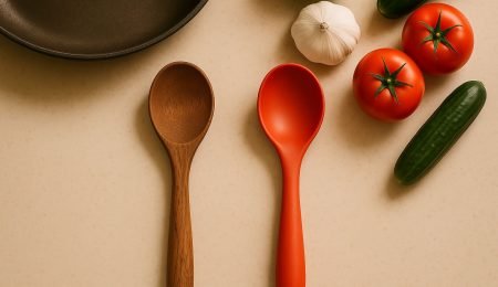 A top-down view of a modern kitchen countertop with a rustic wooden spoon and a colorful silicone spoon side by side. The background includes a nonstick frying pan, fresh vegetables, and a clean, minimal aesthetic. Warm lighting highlights the texture of the wood and the smooth finish of the silicone.